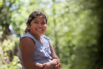 A young latino girl with a smile who is enjoying natures beauty, surrounded by trees with green foliage.