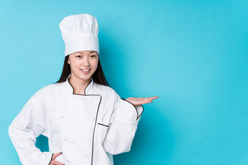 Young chinese chef woman isolated showing a copy space on a palm and holding another hand on waist.