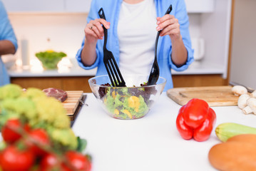 Close up photo of young woman's hands mixing fresh vegetables salad in a glass plate at the kitchen.