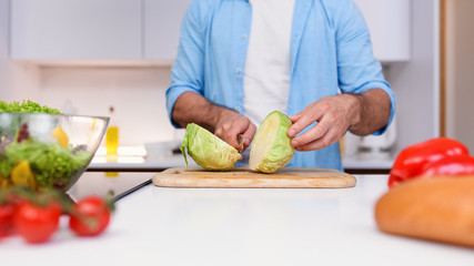 Caucasian man cuts the cabbage with a knife on a wooden board at cozy home kitchen.