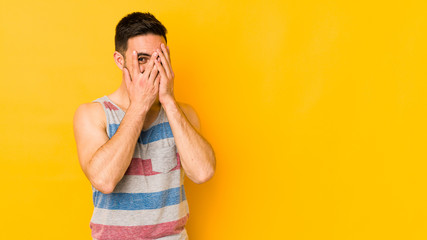 Young caucasian man isolated on yellow bakground blink at the camera through fingers, embarrassed covering face.