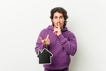 Young man holding a house icon shape keeping a secret or asking for silence.