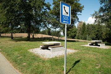 motorway rest area with tables and benches, in the foreground there is a blue s.o.s. sign with a...