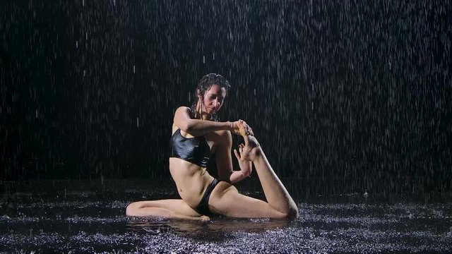 Sports woman doing stretching exercises under the streams of rain. Drops of water fall on body against black background. Studio light creates shimmer on the surface of the water. Slow motion.