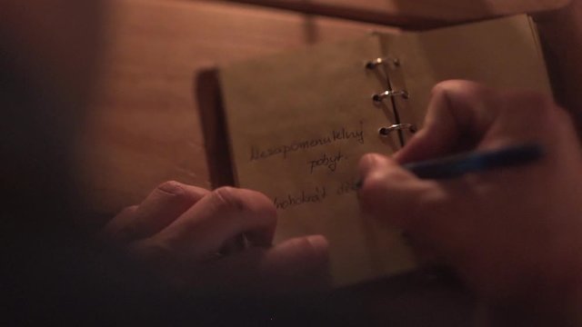 Rack focus close up FHD shot of a man writing a thank you note in a red leather-bound diary ona a wooden table in a dimmed light and closing it afterwards. Man writing an inscription in a notebook.