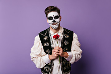 Dark-haired guy with beautiful smile in great mood, posing on isolated background. Photo of Mexican with face art and rose in his hands