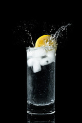 Refreshing glass of water with a lemon slice splashing droplets. Clear glass summer drink with frozen floating ice cubes isolated on black background. Dropping a lemon into a glass