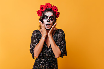 Emotional Mexican dark-haired woman with flowers on her head makes shocked face and looks into camera, touching herself with her hands