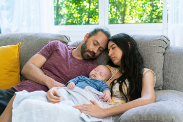 Beautiful Caucasian mother and father with newborn baby son are resting together on sofa in living room. Mom holding sleeping cute infant child boy. Happy Family, parenthood and healthcare concept.