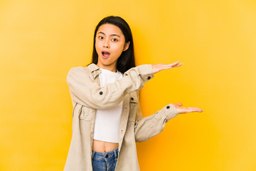 Young chinese woman isolated on a yellow background shocked and amazed holding a copy space between hands.