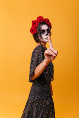 Latin beauty girl with makeup in form of traditional image of skull asks to be silent, showing index finger to camera