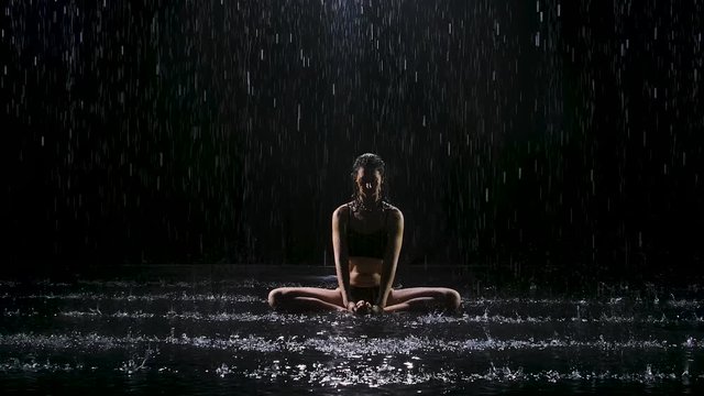 A young woman doing stretching exercises under the streams of rain. Drops of water fall on body against black background. Studio light creates shimmer on the surface of the water. Slow motion.