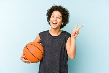 Kid boy playing basketball isolated on blue background joyful and carefree showing a peace symbol with fingers.
