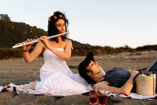 Skilled female musician sitting on blanket on shore and playing flute for girlfriend while resting in evening