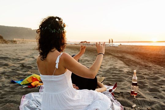 Skilled female musician sitting on blanket on shore and playing flute for girlfriend while resting in evening