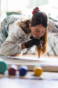 From above of female painter in dirty robe and gloves blowing through thin tube for batik while sitting on wooden floor near colorful abstract painting in art studio