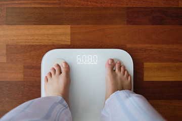 Top view crop faceless barefoot female in cozy pajama standing on digital weight and body fat scales with display showing healthy weight of 60 kg on bathroom floor in morning