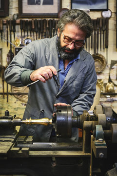 Focused mature bearded goldsmith in eyewear working on polishing machine in workshop while holding pointed metal stick