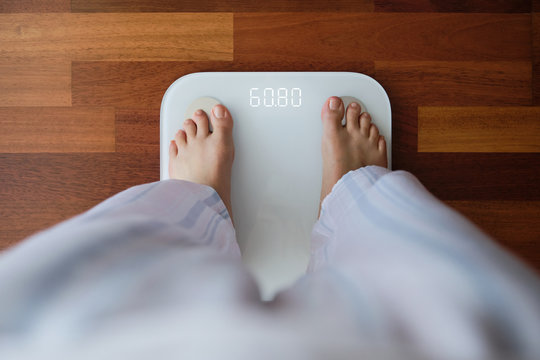 Top view crop faceless barefoot female in cozy pajama standing on digital weight and body fat scales with display showing healthy weight of 60 kg on bathroom floor in morning