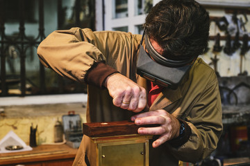 Focused mature goldsmith in head magnifying glasses and uniform working with rectangular engraving in gold frame in workshop near wall with professional tools