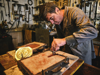 Focused professional mature craftsman cutting sand mold while working with metal casting in craft goldsmith workshop