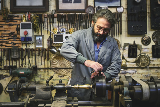 Focused mature bearded goldsmith in eyewear working on polishing machine in workshop while holding pointed metal stick