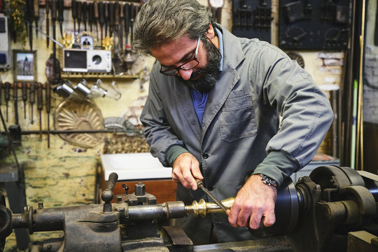 Crop serious mature jeweler in uniform and eyewear sharpening metal stick on professional equipment during work in studio