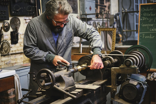 Crop serious mature jeweler in uniform and eyewear sharpening metal stick on professional equipment during work in studio