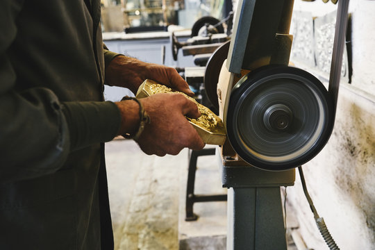Side view of crop unrecognizable male goldsmith polishing edges of metal detail while working with electric grinder in workshop