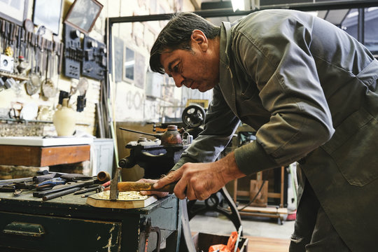 Crop male goldsmith hitting metal detail with hammer during work in workshop