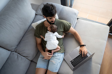 From above male freelancer holding baby while lying on sofa and working on project with netbook at home