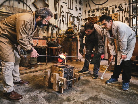 Group of professional male artisans working in weathered workshop and pouring molten metal into mold cavity during metal casting process