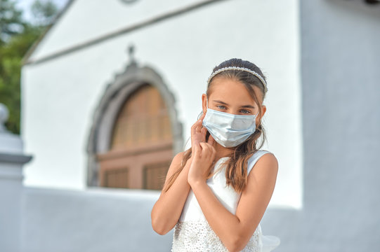 Preteen blond girl in a white dress wearing a protective mask because of the covid-19 pandemic, on the day of her first communion standing outside a church