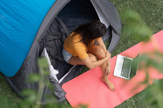From above positive young female in casual clothes sitting on mat with laptop inside tourist tent during camping on green meadow