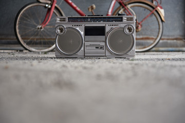 Old fashioned single cassette portable tape player placed on concrete floor against blurred red bicycle and gray shabby wall in garage
