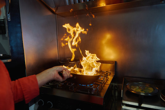 Cropped unrecognizable chef in uniform holding frying pan while flambeing ingredients in frying pan on slow heat standing in restaurant kitchen in evening and looking at bright fire