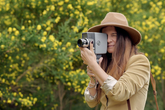 Female photographer in hat with closed eyes filming video with retro video camera while standing near trees with blooming yellow flowers in daylight - Powered by Adobe