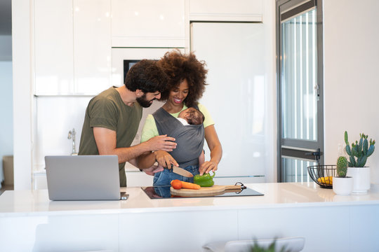 Content Black Mother With Baby In Sling Cutting Vegetables And Arab Father Working On Laptop While Standing At Counter In Kitchen