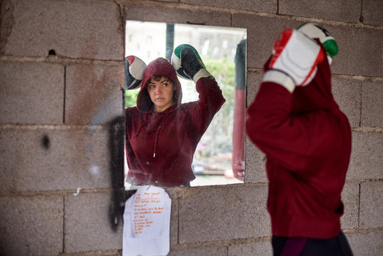 Serious Sportswoman Wearing Hoodie And Boxing Gloves Standing In Double Biceps Pose And Looking In Mirror On Rural Shabby Wall