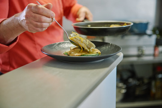 Crop unrecognizable cook standing with frying pan while serving tasty fish fillets on round ceramic plate decorated with lemon zest