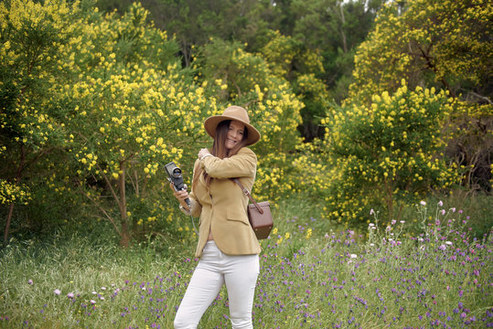 Smiling slim woman in casual wear and hat strolling with old video camera and leather case near bushes with colorful blooming flowers in countryside and looking away