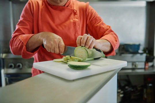 Crop unrecognizable chef in uniform cutting fresh zucchini into thin slices using knife and cutting board during cooking process in restaurant