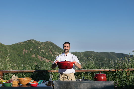 Professional cook in uniform standing with saucepan in the hands against mountainous terrain on sunny day