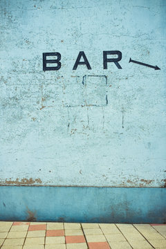 Old blue cement wall with scratches on surface and BAR inscription with painted arrow near tiled floor in daylight