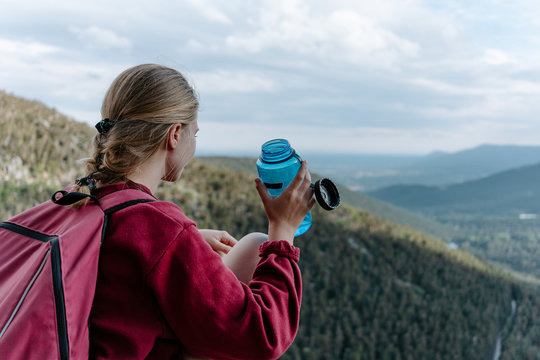 Side view of young female backpacker sitting on stone against green forested hill and drinking water while resting during hiking in Navacerrada in Spain