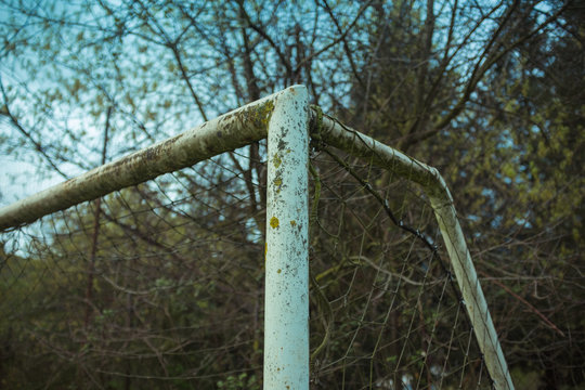 From below of football goal with metal beams with shabby corroded surface and grid near overgrown trees with thin branches under blue sky
