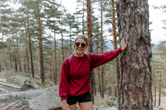 Young female hiker in sunglasses standing leaning on tree trunk and looking at camera during trekking in forest in Navacerrada in Spain