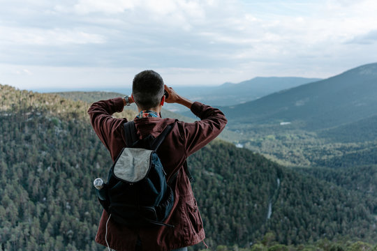 Back view of unrecognizable male tourist with backpack standing on mountain slope and admiring view of green hills while observing landscape with binoculars in Navacerrada in Spain