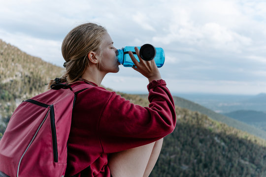 Side view of young female backpacker sitting on stone against green forested hill and drinking water while resting during hiking in Navacerrada in Spain
