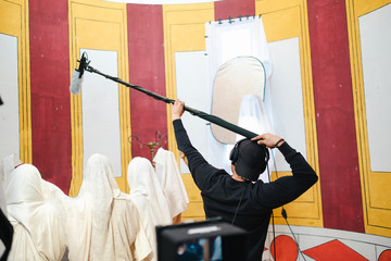 Back view faceless assistant raising professional shotgun microphone above group of Arabs in white national clothes while working in creative studio during video recording